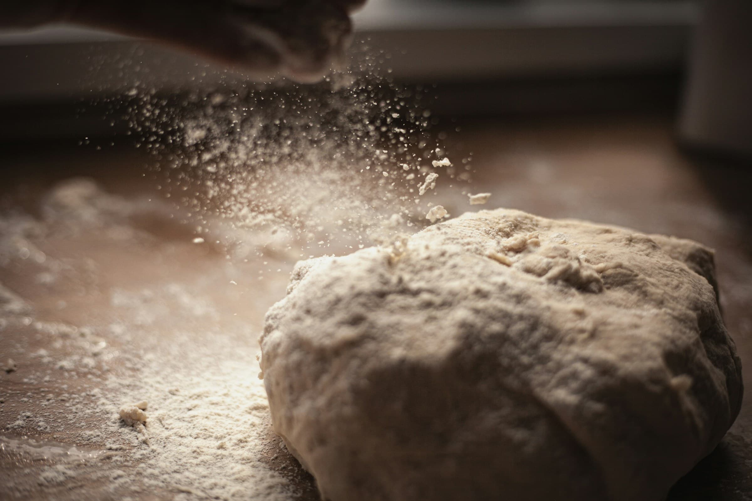 Hands dusting flour onto a dough ball on a dark wood surface